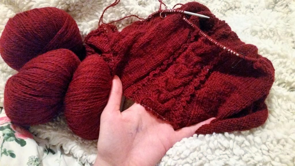 A work in progress in red fingering weight yarn is being held in the knitter's hand on top of a floral bedspread and a fuzzy blanket.