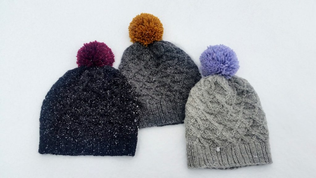 Three grey knit hats with an argyle cable pattern and colourful pom-poms lying in the snow