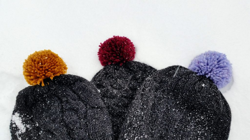 Three dark grey hats with varying knit textures and colourful pom-poms are lying in the snow.