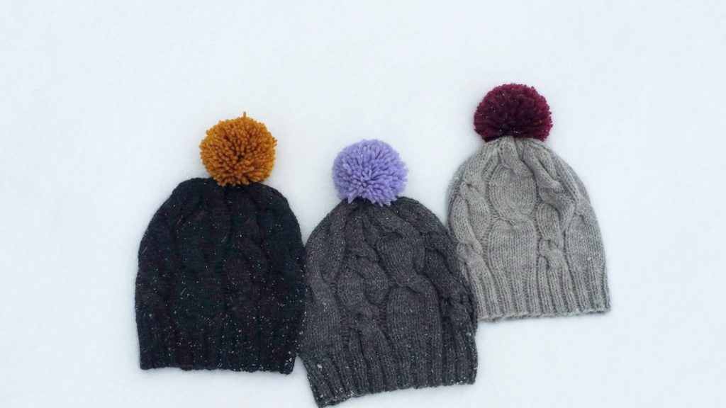 Three grey knit hats with alternating cable patterns and colourful pom-poms are lying in the snow.