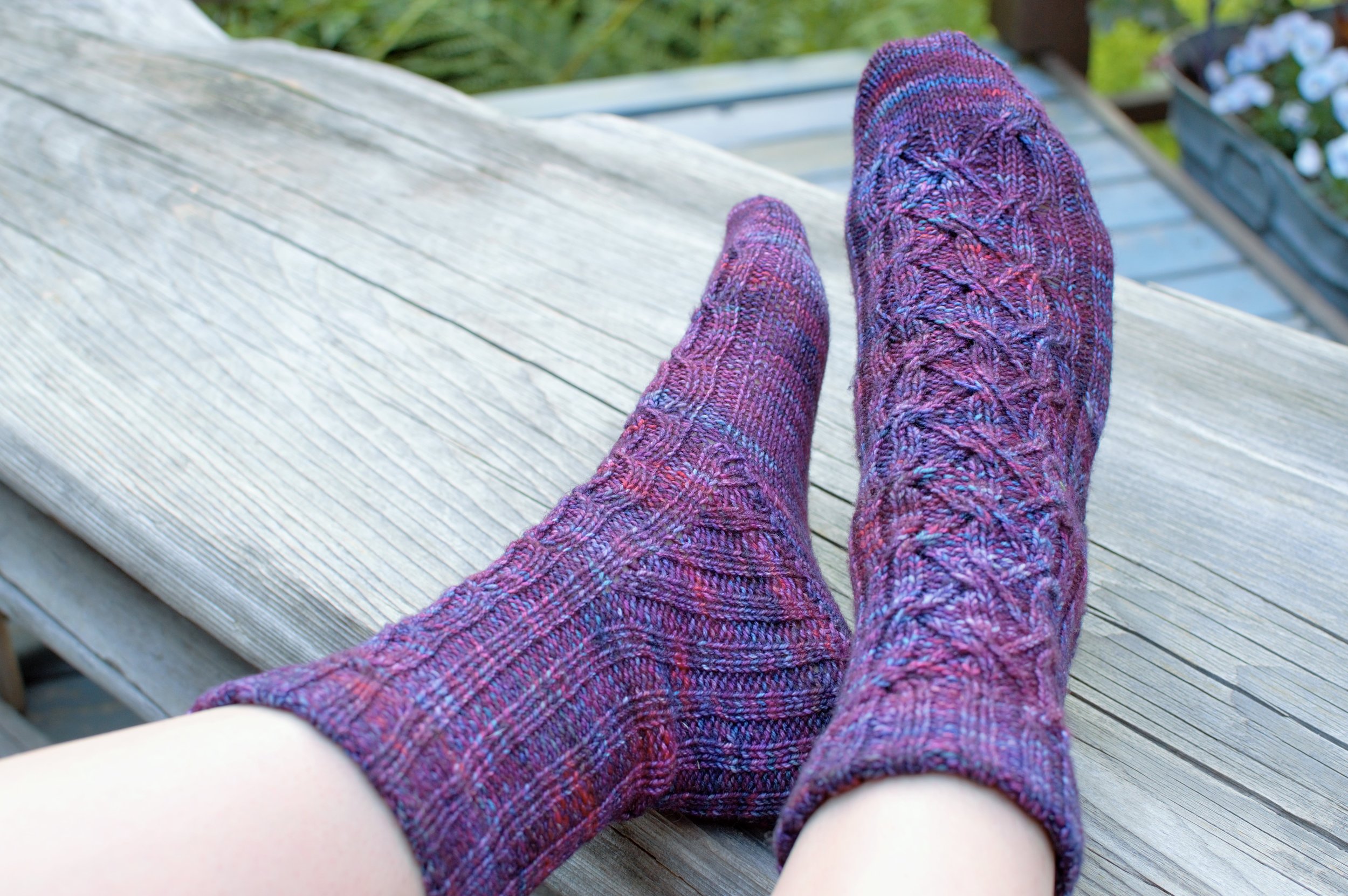 A pair of feet wearing variegated purple socks with cables up the front and ribbing along the back are resting on a driftwood bench.