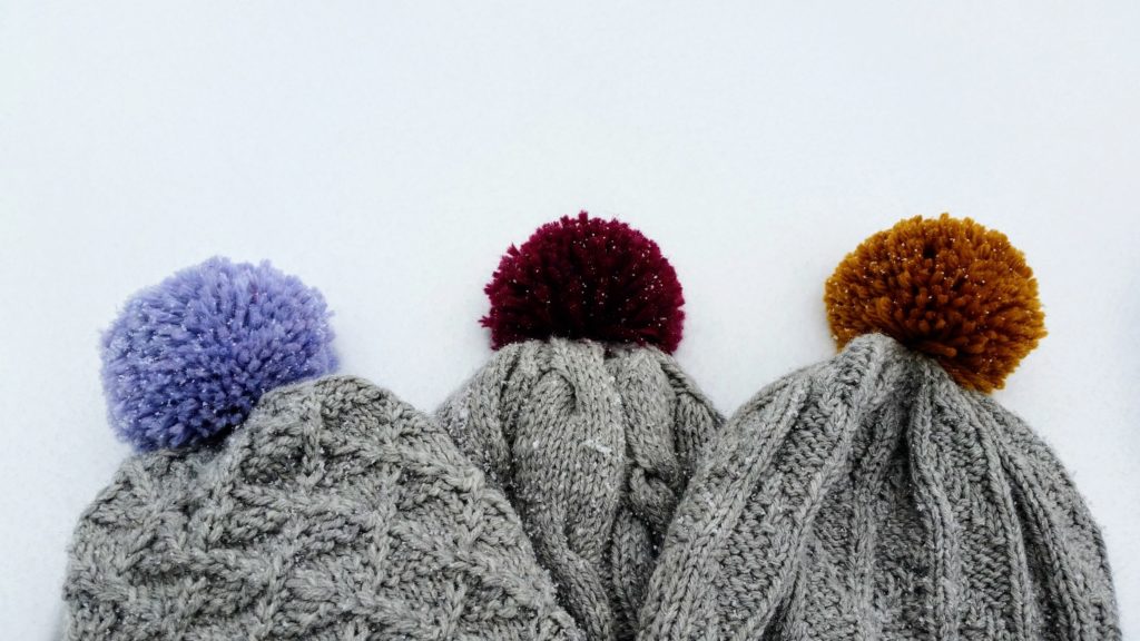 Three light grey hats with varying knit textures and colourful pom-poms are lying in the snow.