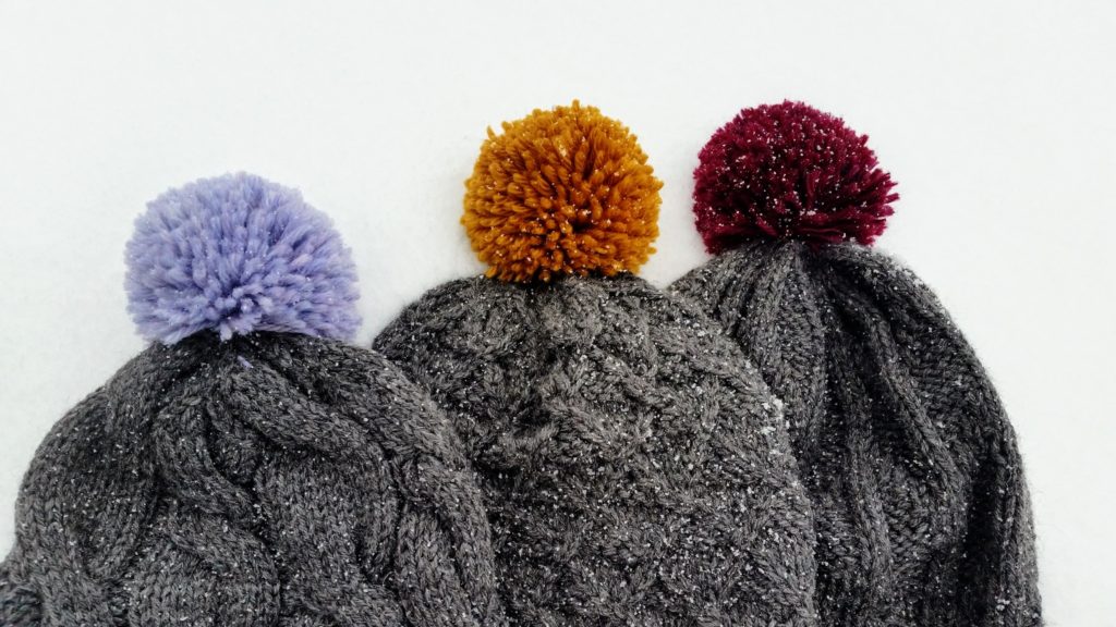 Three charcoal grey hats with varying knit textures and colourful pom-poms are lying in the snow.