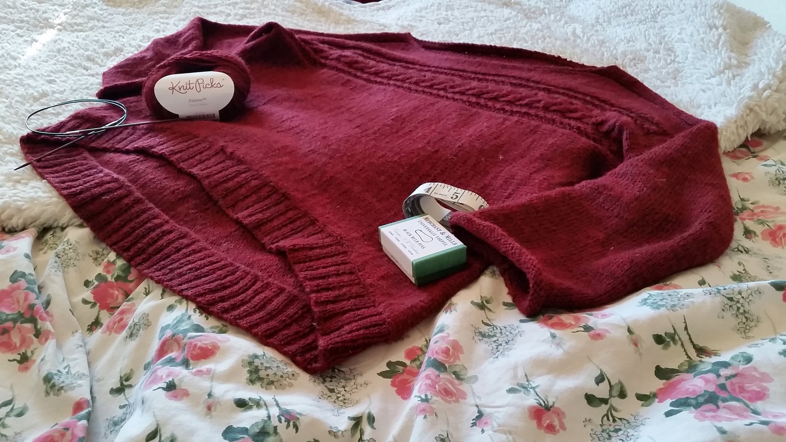 A red knit pullover with a cable that runs horizontally across the chest is laid flat against a floral bedspread and a fuzzy blanket.
