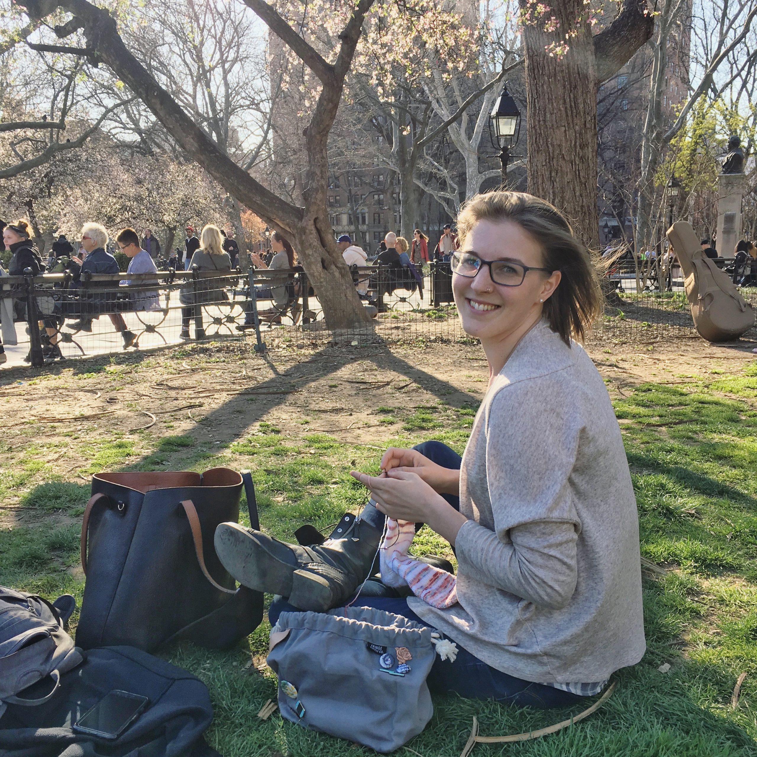 A blonde woman sitting cross-legged in a grassy area of Washington Square Park, knitting a pair of pink socks.