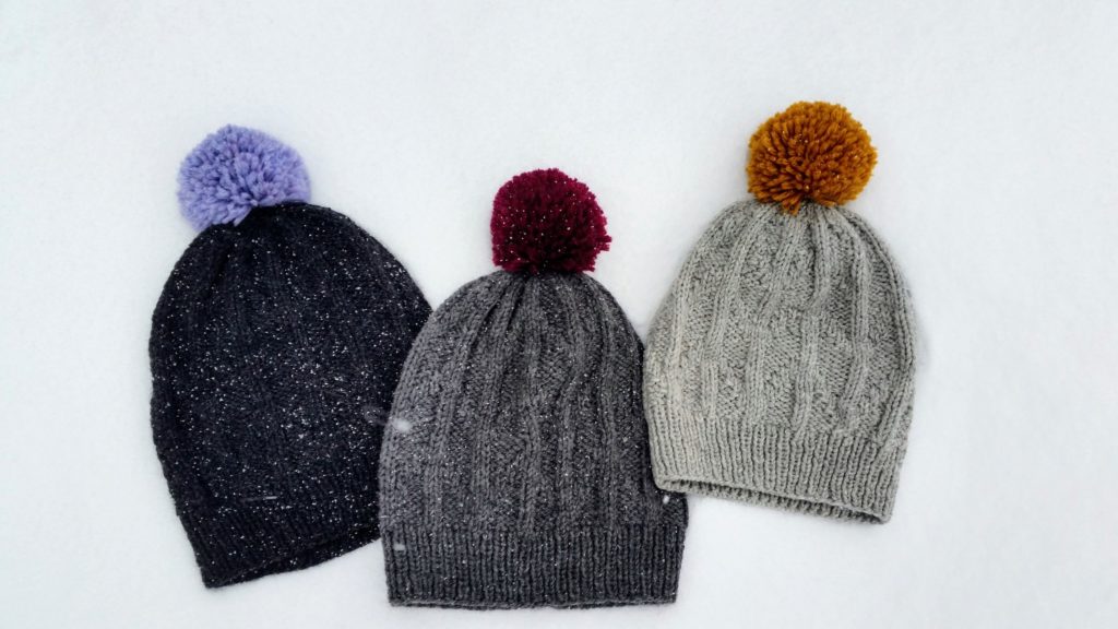 Three grey knit hats with ribbing that resembles a chevron pattern and colourful pom-poms are lying in the snow.